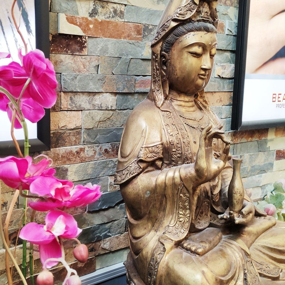 Bronzefarbene Buddhastatue vor einer Steinwand mit pinken Orchideen